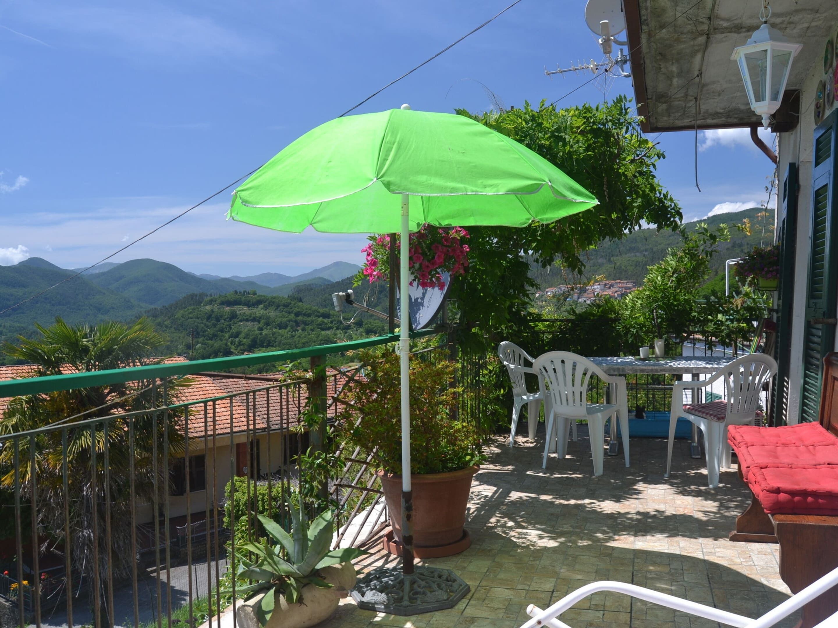 Un balcone con un ombrello verde, sedie bianche, una panca imbottita rossa e piante in vaso.