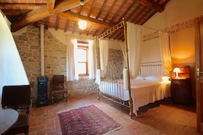 Un dormitorio con pared de piedra, cama con dosel, una silla de madera, una mesa y una lámpara.