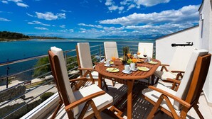 Une table ronde en bois, entourée de chaises rembourrées blanches, est placée sur un balcon qui offre une vue sur la côte, avec un ciel dégagé et quelques nuages épars.