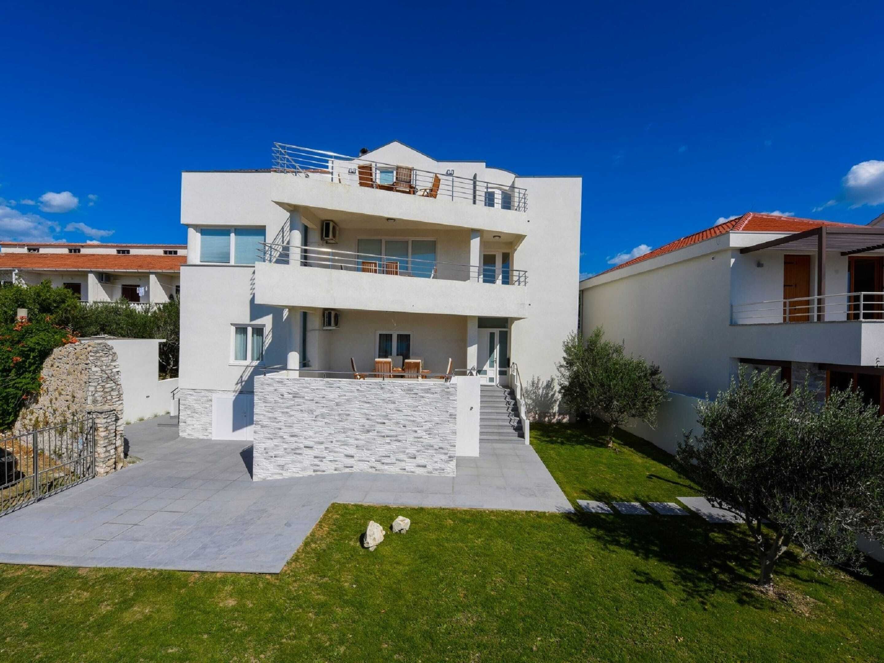 A modern two-story building with a flat roof, a stone retaining wall, and a paved courtyard with a small lawn area.