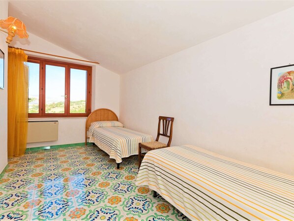 A room with two single beds, a chair, a window with curtains, a radiator, and a colorful tiled floor.