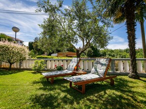 Two wooden lounge chairs with patterned cushions, a small table with a drink and flowers, and a white fence with a palm tree and shrubbery in the background. - Lovely Apartment With Terrace & Garden Grill (Malinska-Dubasnica)