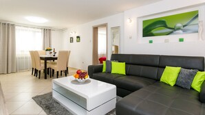 A modern living room with a black leather sofa, a white coffee table, and a dining area with a table and chairs. - Lovely Apartment With Terrace & Garden Grill (Malinska-Dubasnica)