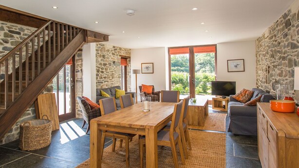 A spacious living area with a wooden dining table, comfortable seating, a stone wall, and a staircase. - Holiday Home in Tavistock With Garden (Tavistock)