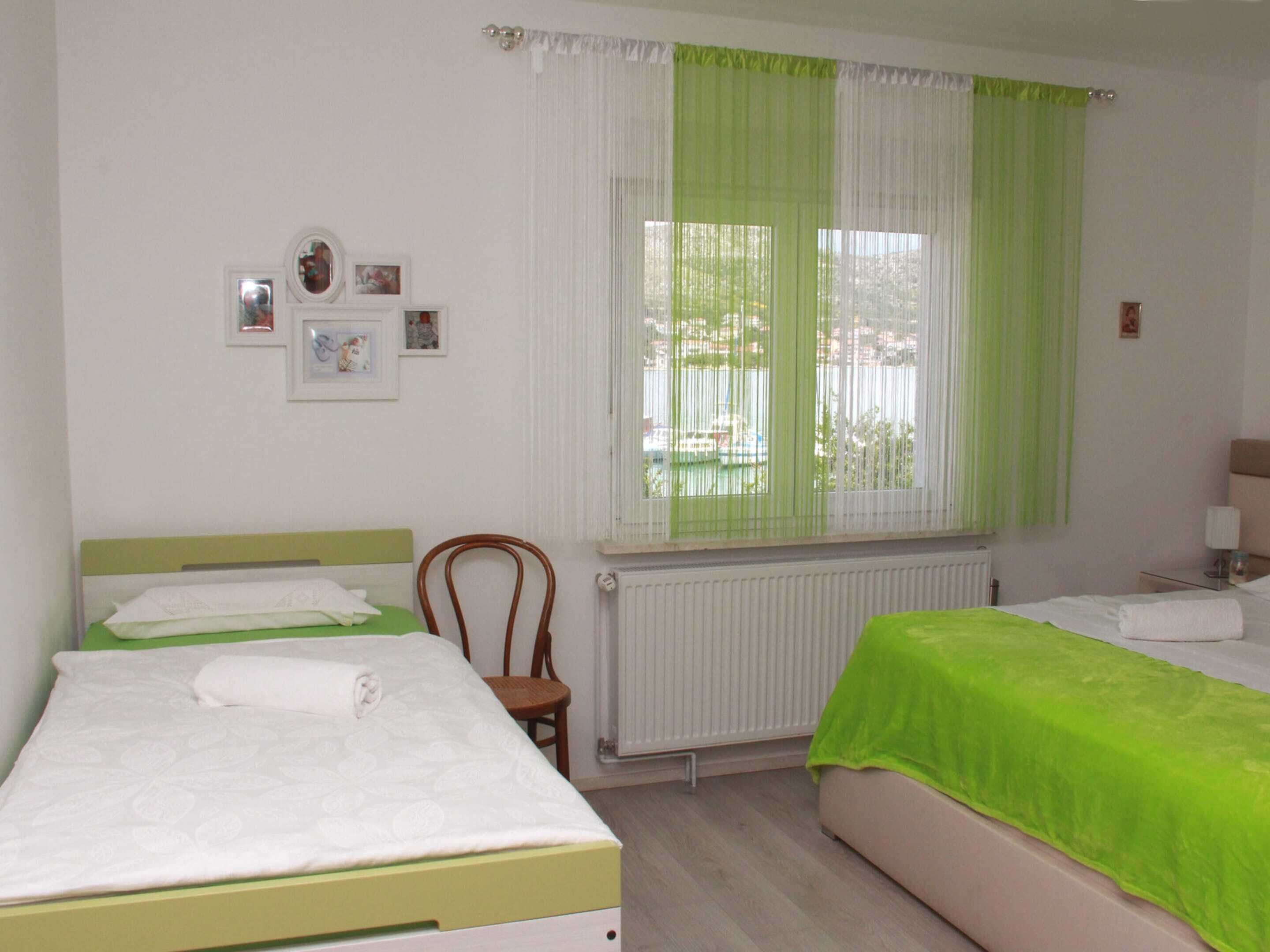 A bedroom with two beds, a chair, a radiator, and a window with green curtains.