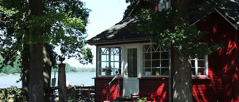 A red wooden house with a white door and windows, surrounded by trees and a body of water.