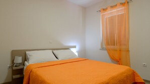 Un dormitorio con cama, mesita de noche, lámpara y ventana con cortinas translúcidas.