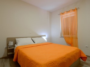 Une chambre à coucher avec un lit, une table de chevet, une lampe et une fenêtre avec des rideaux légers.