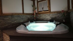 A hot tub in a room with stone walls, wooden beams, and a window with a curtain.