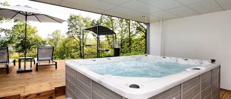 A hot tub on a wooden deck with a pergola and chairs, surrounded by trees.