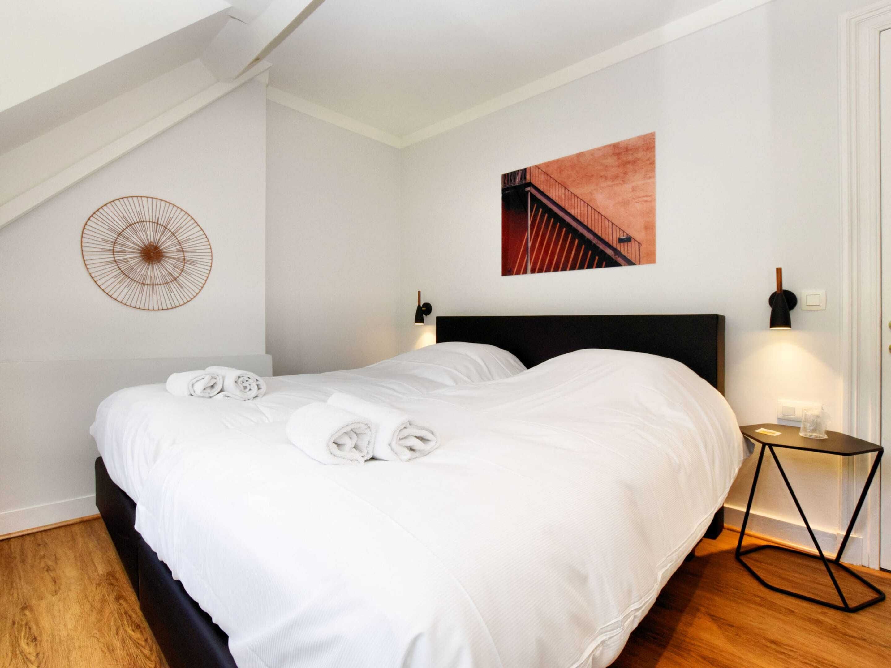 A neatly made bed with rolled towels, a modern headboard, a wall-mounted light fixture, and a framed picture on the wall.