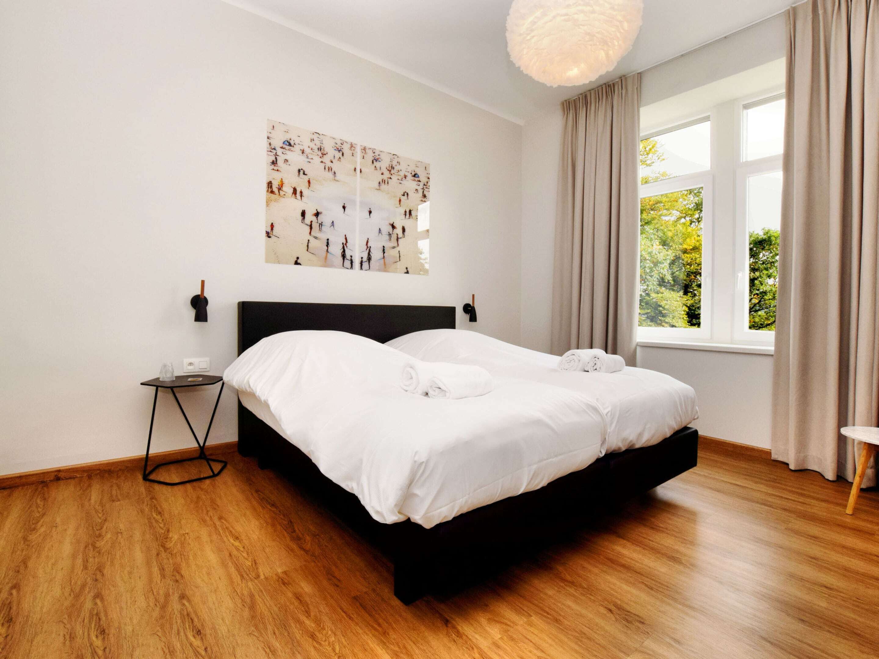 A modern hotel room with a large bed, a side table, a wall-mounted light fixture, and a window with curtains.