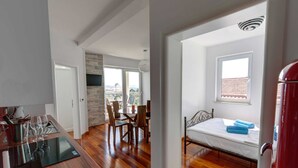A modern room with a wooden floor, a bed with a white mattress, a dining table with chairs, and a kitchen area with a stove and wine glasses. - Adorable Apartment in Murter with Hot Tub (Murter-Kornati)