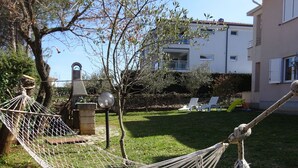 A hammock in a backyard with a lawn, a barbecue grill, and a building in the background. - Large Apartment in Malinska near Sea (Malinska-Dubasnica)