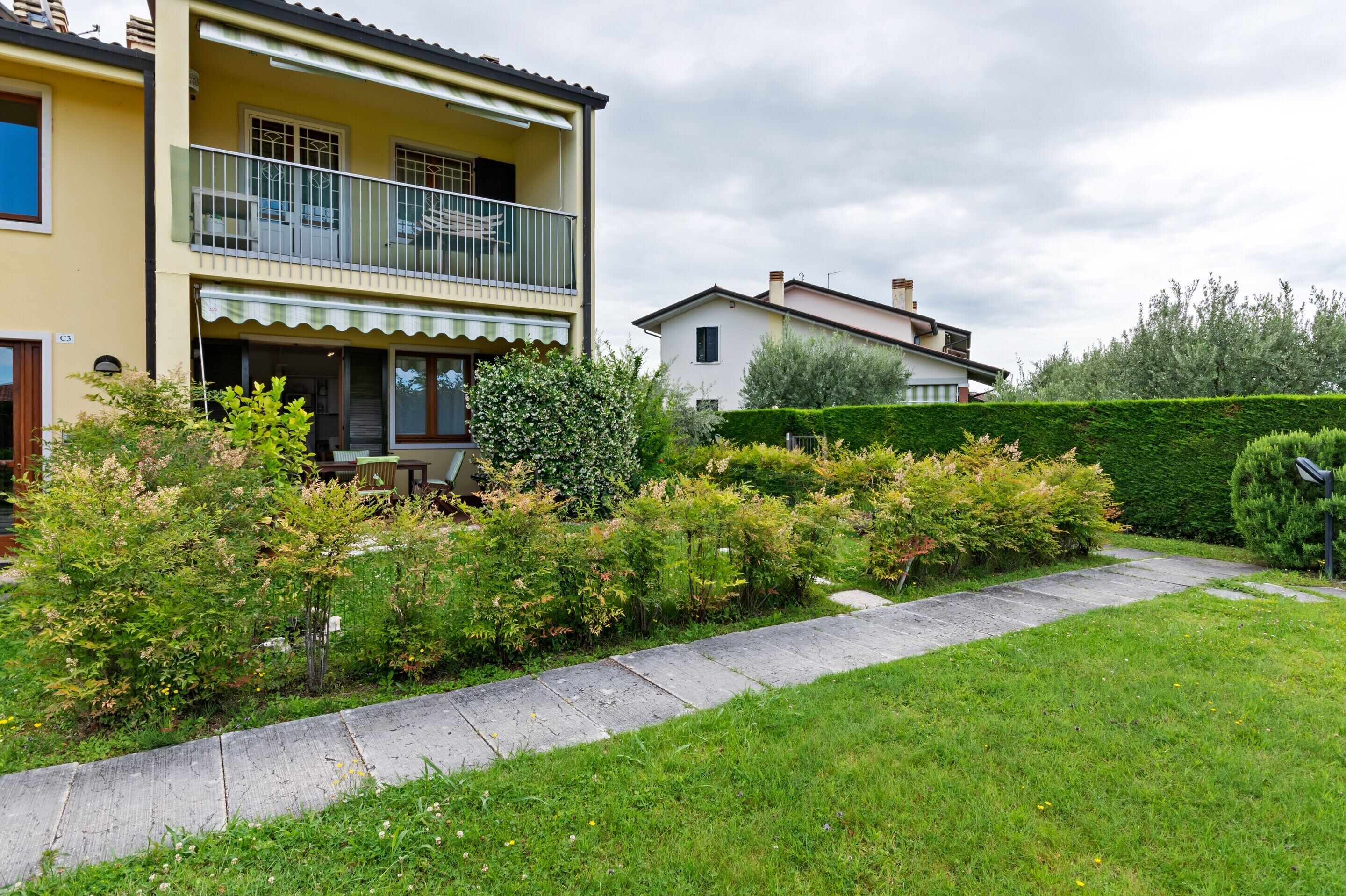 Un immeuble résidentiel de deux étages doté d’un balcon, d’un jardin avec différentes espèces de plantes et d’un ciel dégagé.