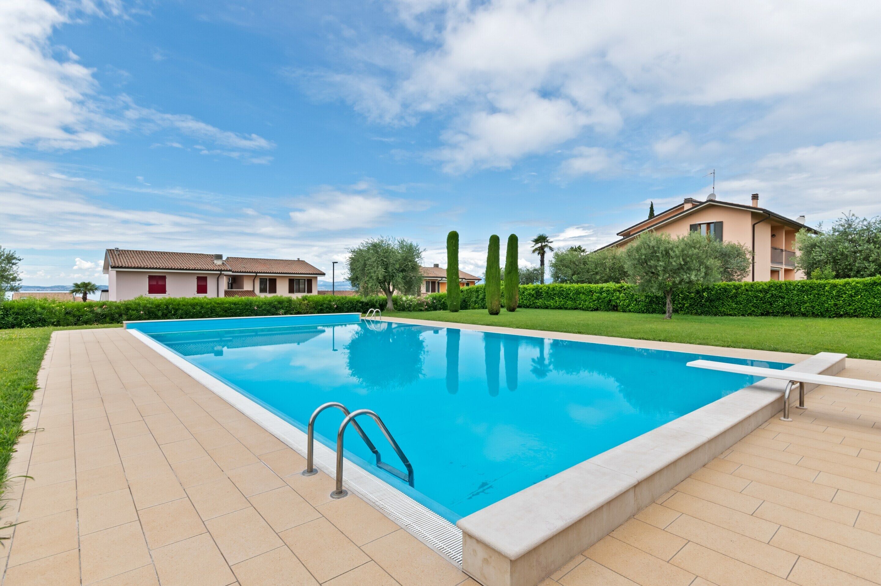 A swimming pool with a tiled deck, surrounded by greenery and two buildings.