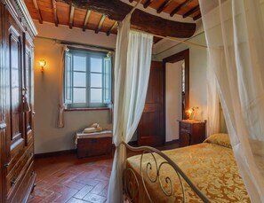 A bedroom with a four-poster bed, a wooden wardrobe, a bedside table with a lamp, a window with curtains, and a chest of drawers. - Luxury Pool Villa in Cortona (Cortona)
