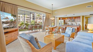 A spacious living room with a large TV, comfortable seating, and a view of a balcony with outdoor furniture. - Maui Resort Rentals: Honua Kai Hokulani 109 – Ground Floor Interior Courtyard 2BR, Steps to Beach/Pools, Large Lawn Area, Partial Ocean Views! (Lahaina)