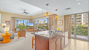 A modern kitchen with a central island, a dining area with a sofa and chairs, a ceiling fan, and large windows with curtains. - Maui Resort Rentals: Honua Kai Hokulani 109 – Ground Floor Interior Courtyard 2BR, Steps to Beach/Pools, Large Lawn Area, Partial Ocean Views! (Lahaina)