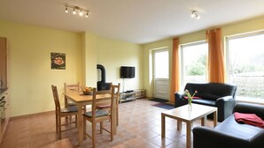 A living room with a wooden dining table, chairs, a black sofa, a small coffee table, a TV, and a window with curtains. - Ferienwohnung in Zierow (Zierow)