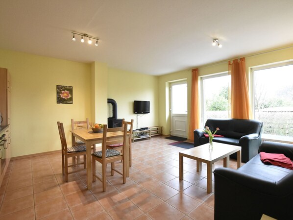 A living room with a wooden dining table, chairs, a black sofa, a small coffee table, a TV, and a window with curtains. - Ferienwohnung in Zierow (Zierow)