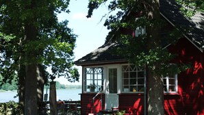 Ett rött trähus med en vit dörr och vita fönster, omgivet av träd och en vattenyta.