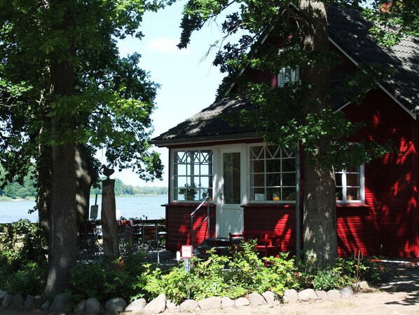A red wooden house with a white door and windows, surrounded by trees and a body of water. - Charming Coastal Retreat in Zierow (Zierow)