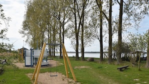 A playground with a swing set and a play structure, surrounded by trees and a body of water. - Modernes Ferienhaus in Boiensdorf am Meer (Boiensdorf)