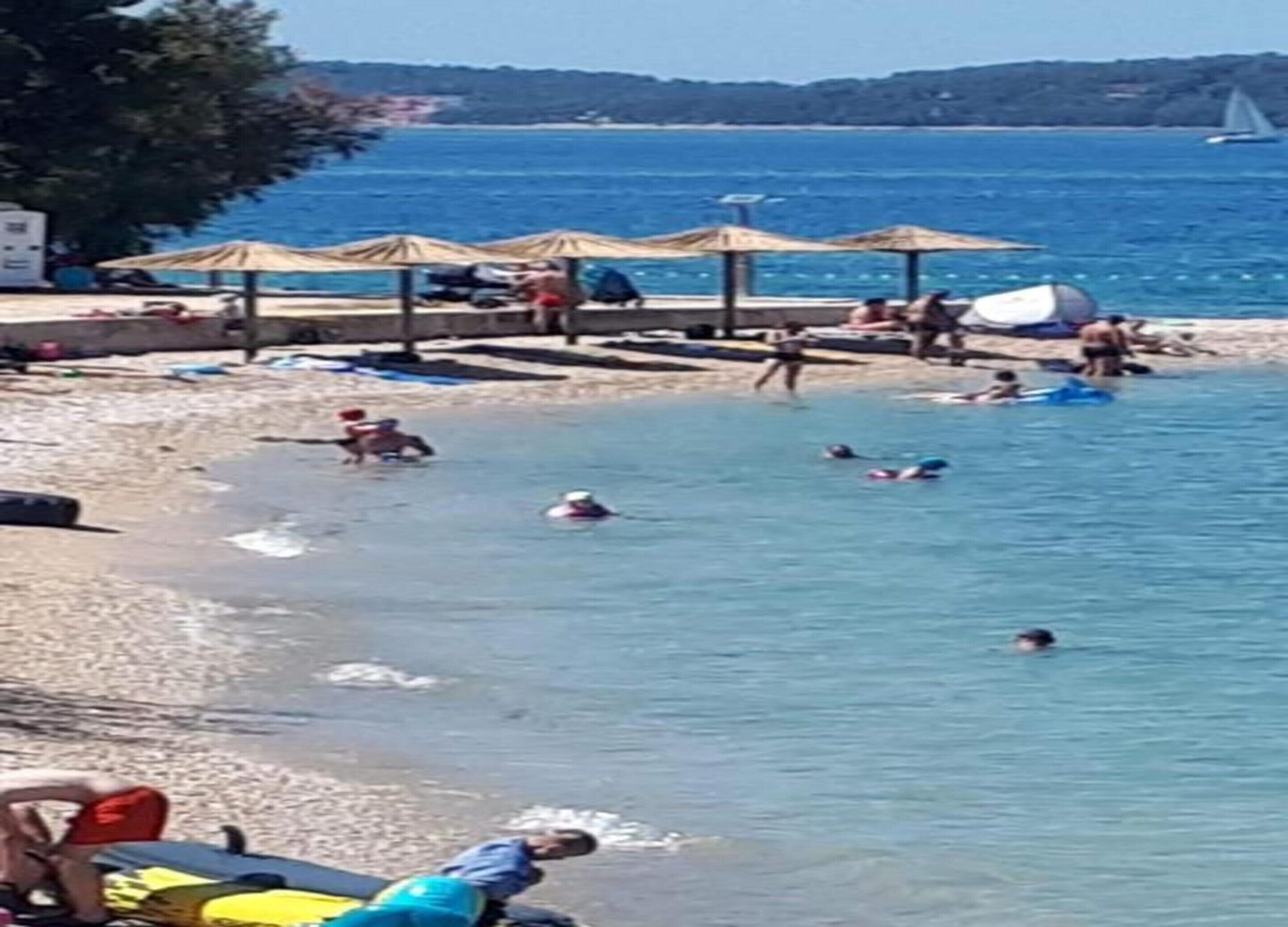 Una playa con sombrillas, gente nadando y barcos al fondo.