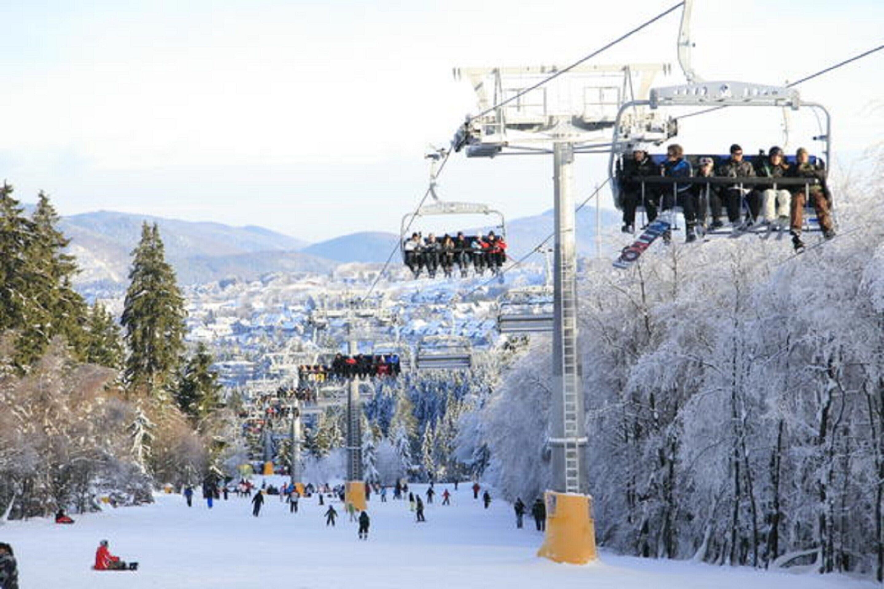 A ski lift with passengers, surrounded by snow-covered trees and a town in the background.