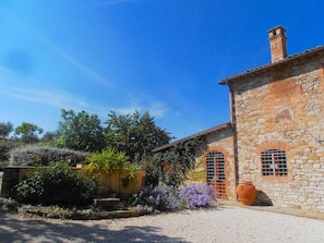 Un bâtiment en pierre avec un jardin, un grand pot en terre cuite et un ciel d’un bleu limpide.