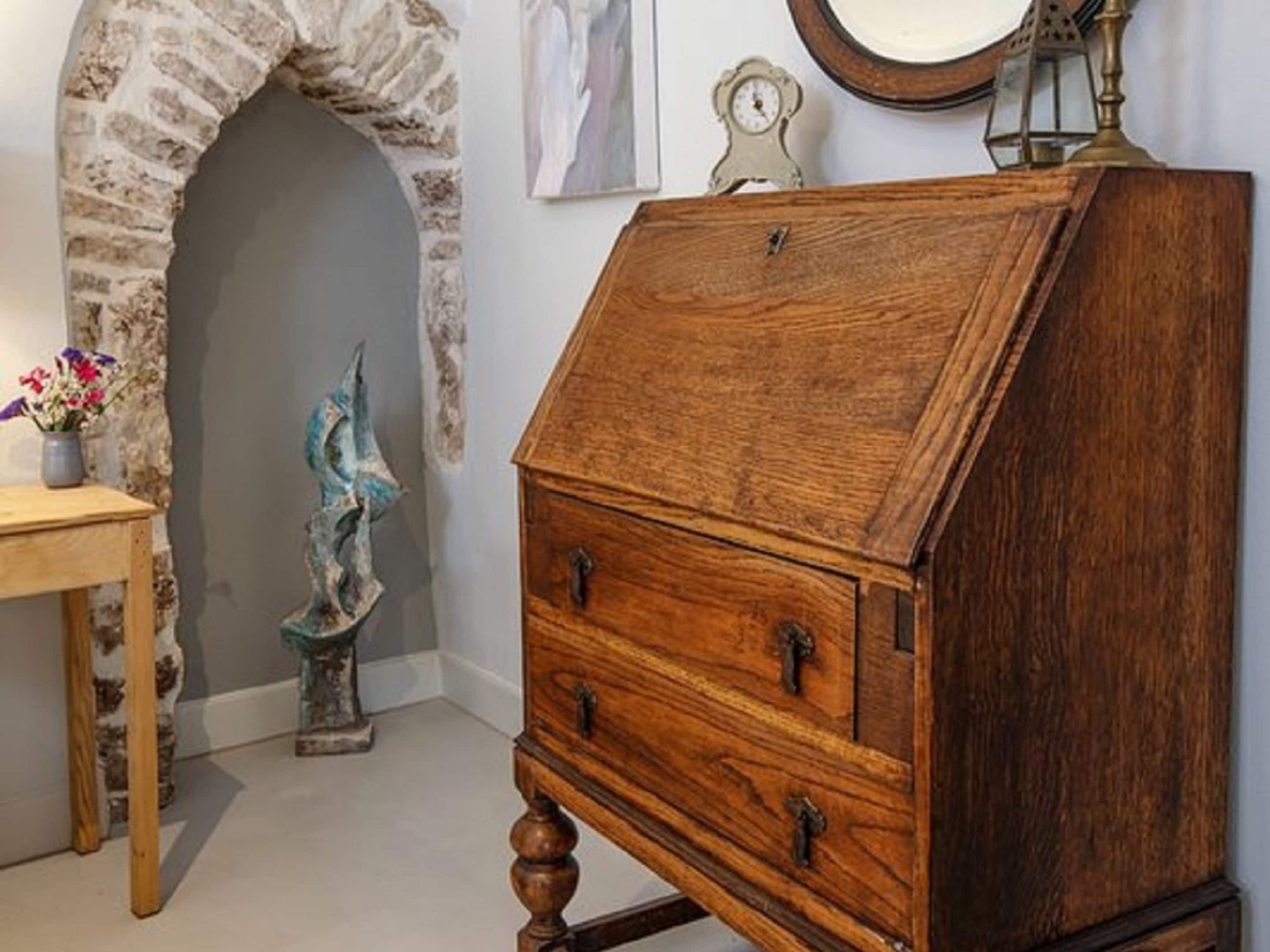 A wooden chest of drawers with a slatted top, a stone archway, a small table with a vase of flowers, and a wall-mounted clock.