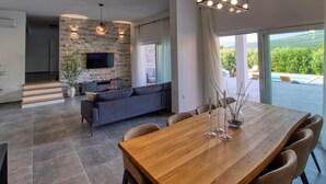 A modern living room with a stone wall, a flat-screen TV, a wooden dining table, and a large window overlooking a pool. - Villa With Private Pool and Garden (Novigrad)