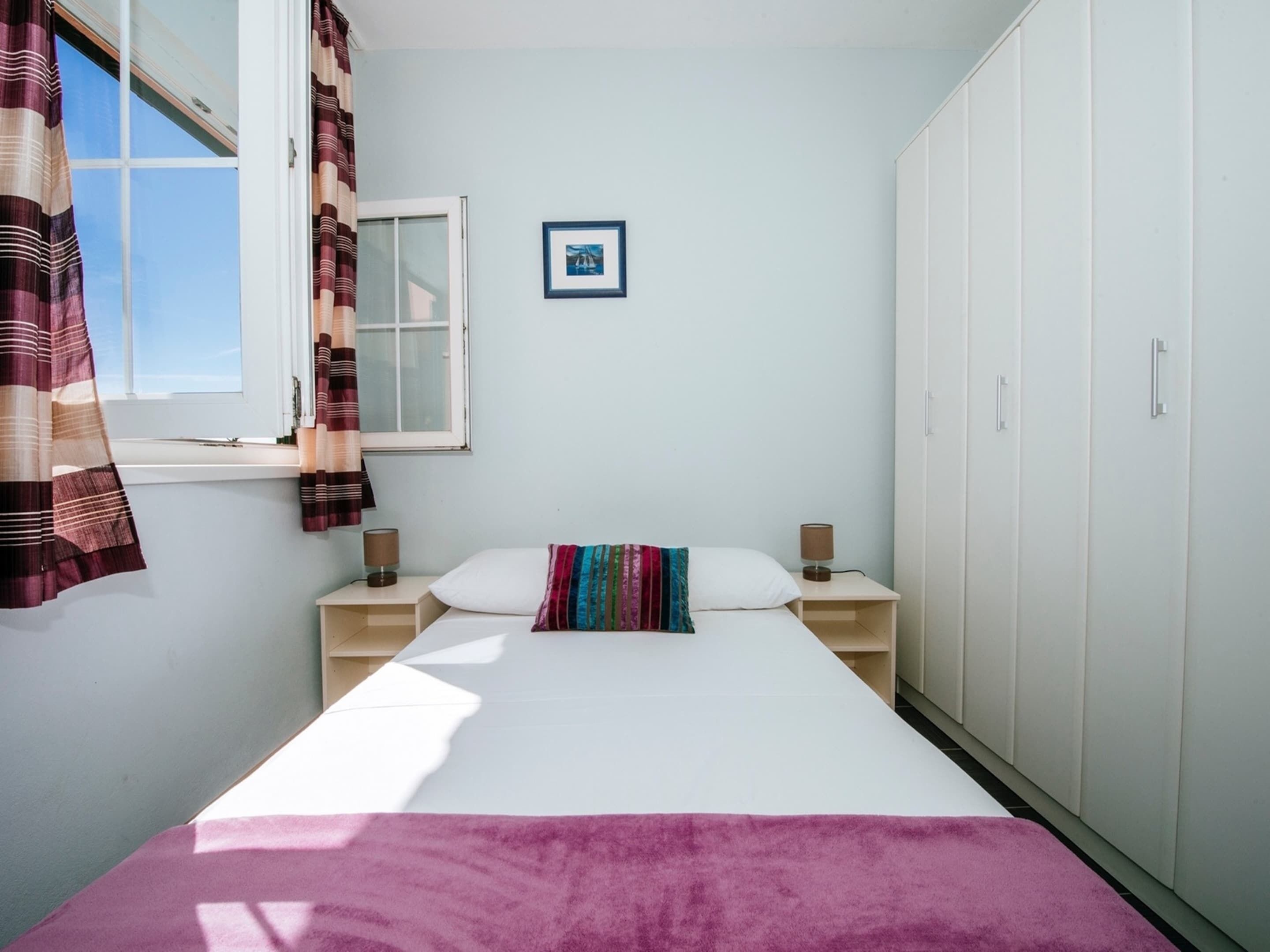 A bedroom with a bed, two bedside tables, a wardrobe, a window with curtains, and a framed picture on the wall.