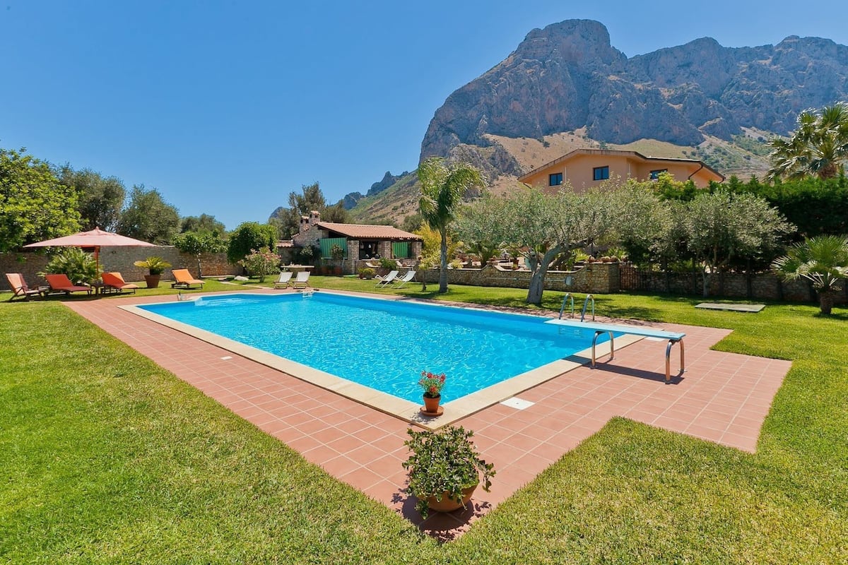 A swimming pool with a mountain backdrop, surrounded by a well-maintained lawn and a few lounge chairs.