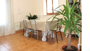Un comedor con una mesa preparada para cuatro personas, rodeada de sillas, una planta en maceta y una estantería al fondo.