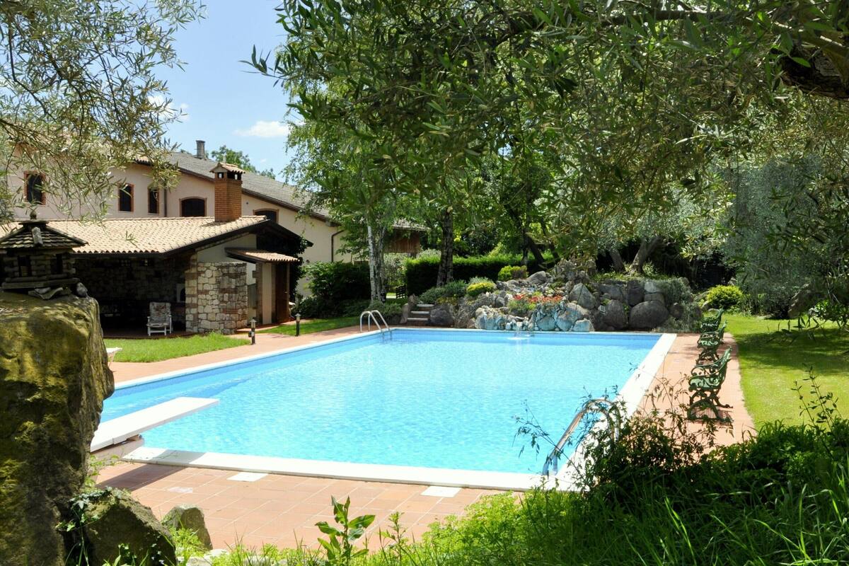 Una piscina rectangular con solárium de baldosas, rodeada de abundante vegetación y un edificio de piedra con tejado de tejas.
