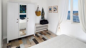 Une chambre à coucher avec un lit, une armoire, un banc, une fenêtre avec des rideaux et une vue sur la mer.