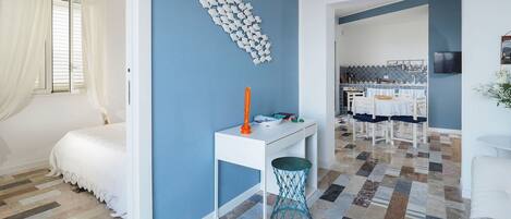 Une pièce avec un mur d’accent bleu, un lit blanc, une petite table blanche surmontée d’un panier bleu, et un sol carrelé aux motifs variés.