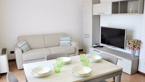 A modern living room with a sofa, a dining table with plates and glasses, a flat-screen TV, and a small plant. - Holiday Home in Lazise Near Lake Garda (Lazise)