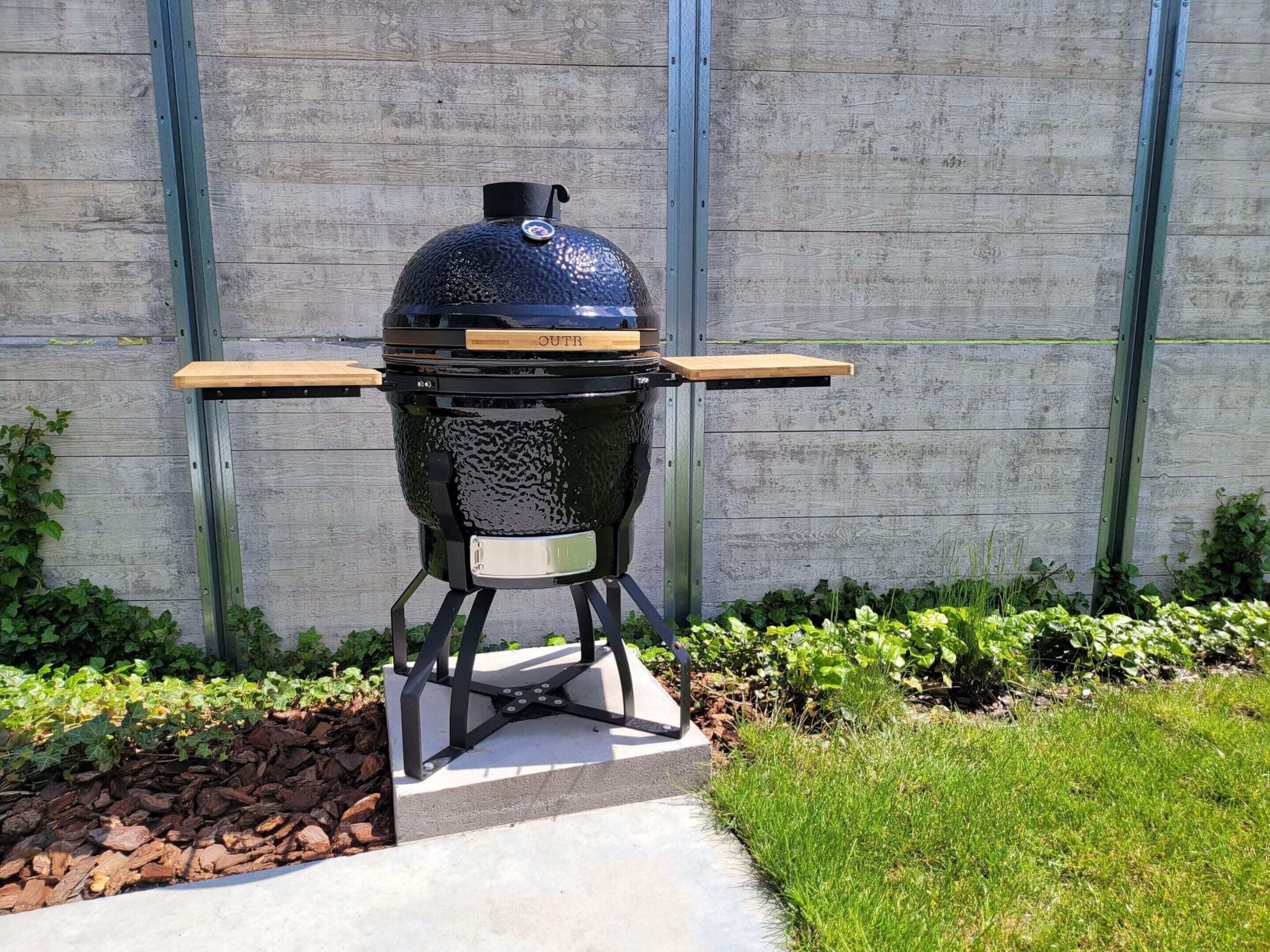 A black outdoor grill with a wooden lid and two side grates, mounted on a concrete base with a metal frame, set against a wooden fence.