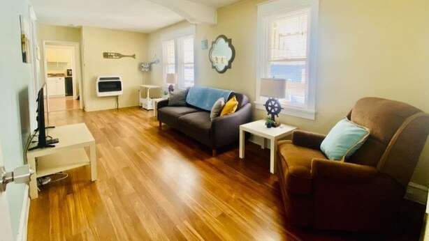 Amplia sala de estar con suelo de madera, un sofá gris, un sillón marrón y una pequeña mesa blanca.