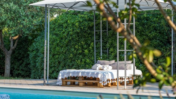Un lit installé au bord de la piscine, surmonté d’un dais, entouré d’une haie verdoyante et donnant sur une piscine à l’eau cristalline.
