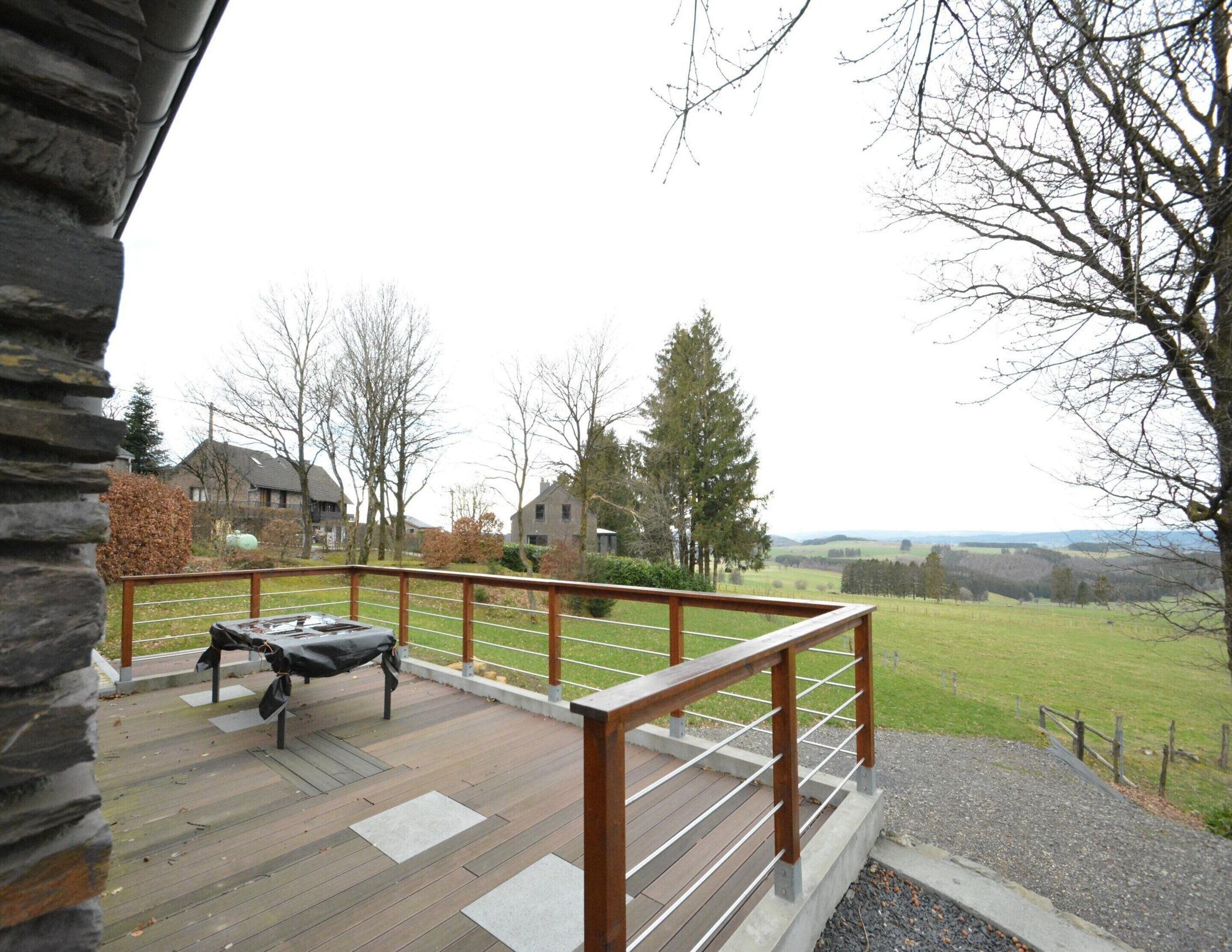A wooden deck with a metal railing, a covered grill, and a view of houses and trees in the background.