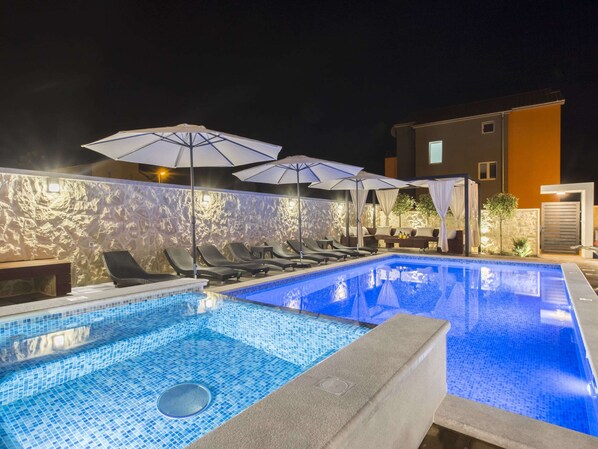 A swimming pool with lounge chairs and umbrellas, surrounded by a textured wall and a building with a door and windows. - Exquisite Villa with Private Hot Tub in Grebaštica (Sibenik)