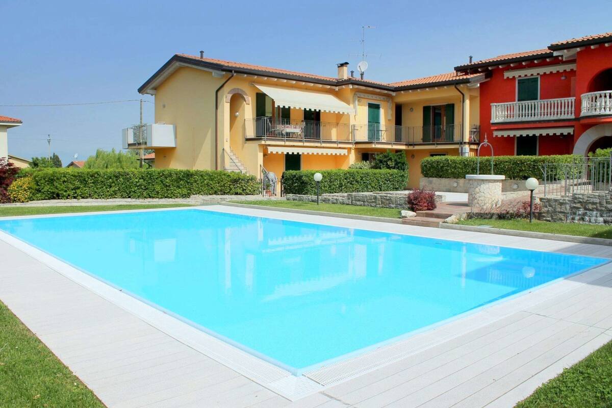Une grande piscine avec une terrasse blanche, un bâtiment jaune doté de balcons et un bâtiment rouge avec un balcon.