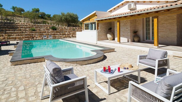 A pool area with lounge chairs and a table, a stone wall, and a building with a tiled roof. - Belvilla by OYO Villa Kika (Noto)