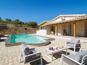 A pool area with lounge chairs and a table, a stone wall, and a building with a tiled roof. - Belvilla by OYO Villa Kika (Noto)