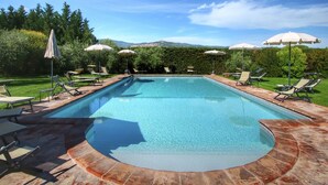 Ein rechteckiger Pool mit gefliestem Rand, umgeben von Liegestühlen und Sonnenschirmen, vor einer Kulisse aus Grünpflanzen und sanften Hügeln.