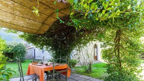 Eine Terrasse im Freien mit Esstisch, umgeben von Grünpflanzen und einem Steinhaus im Hintergrund.
